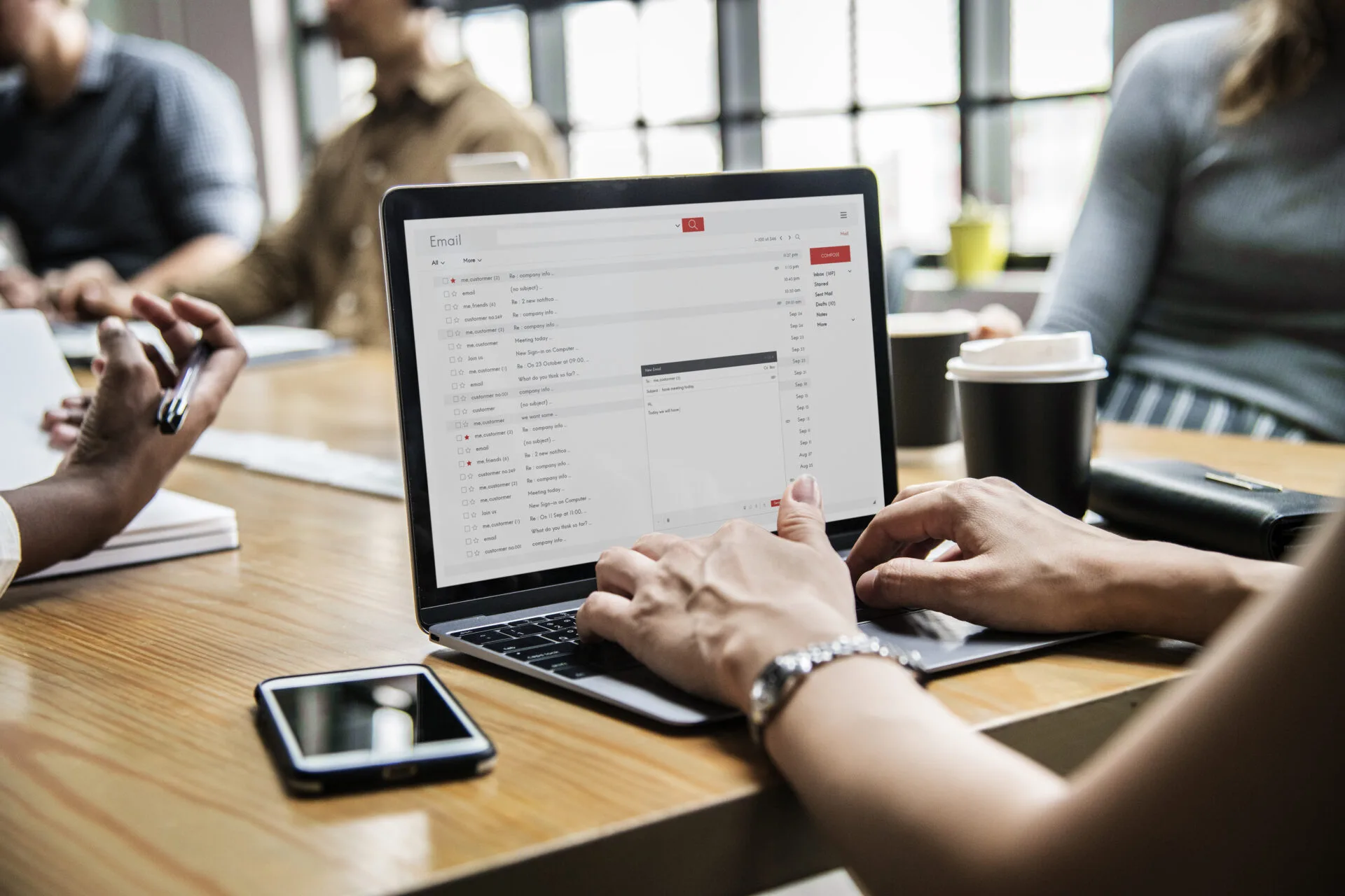 Person using a laptop with email open at a meeting table shared with colleagues