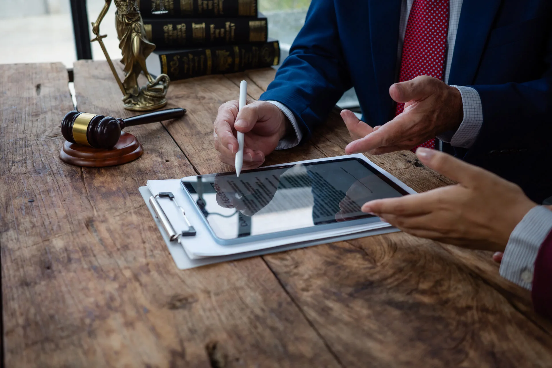 Professionals in suits reviewing a document on a tablet, at a table with a judge's gavel and a statue of justice