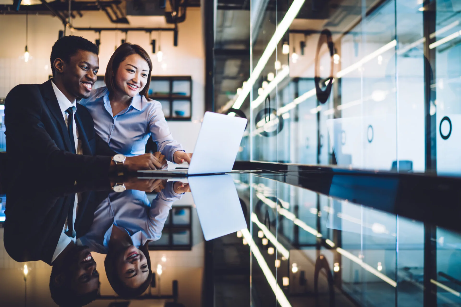 Two smiling professionals looking at a laptop in a modern office with glass walls