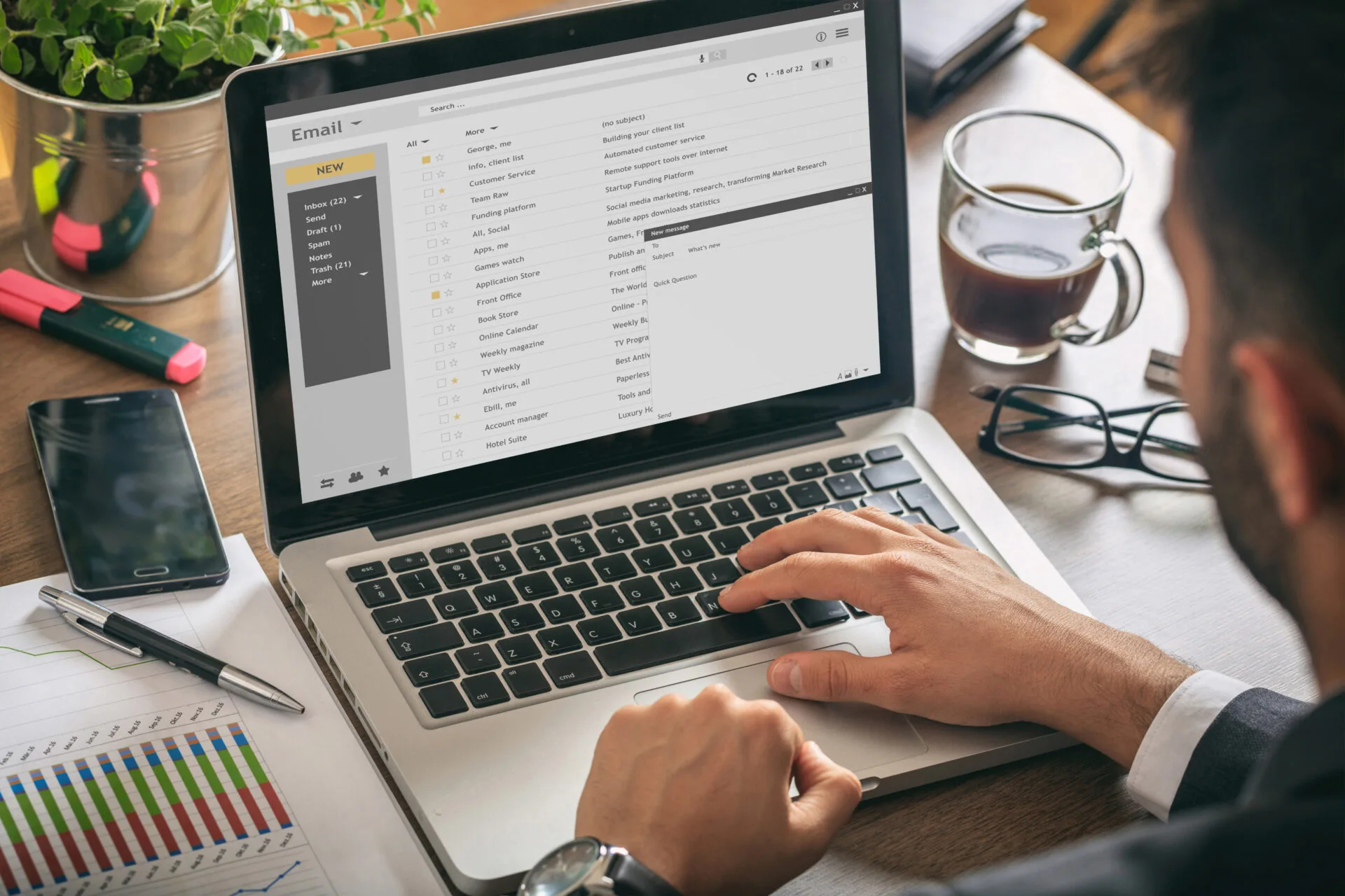 Man typing on a laptop with the email inbox open, at a desk with coffee and a phone