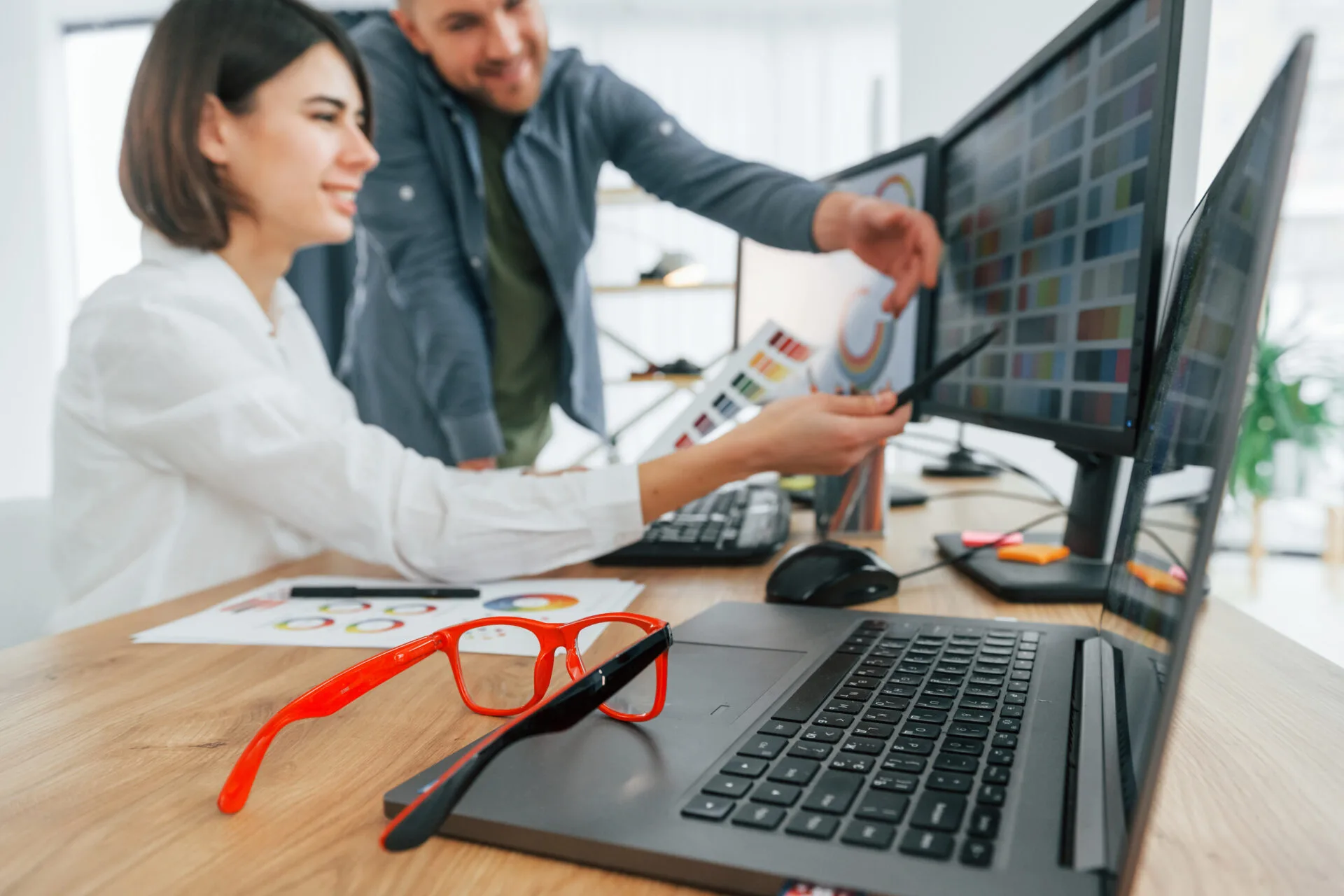 Two smiling designers reviewing colour palettes on monitors, with red glasses in the foreground