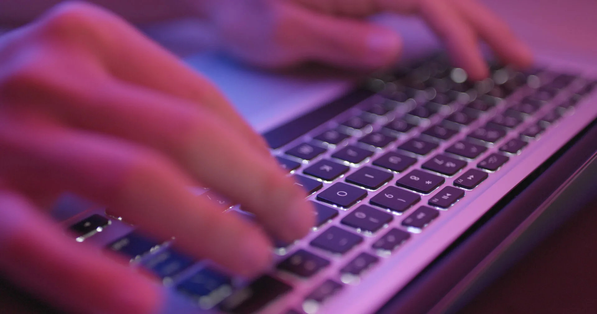 Hands typing on a laptop keyboard lit by pink light
