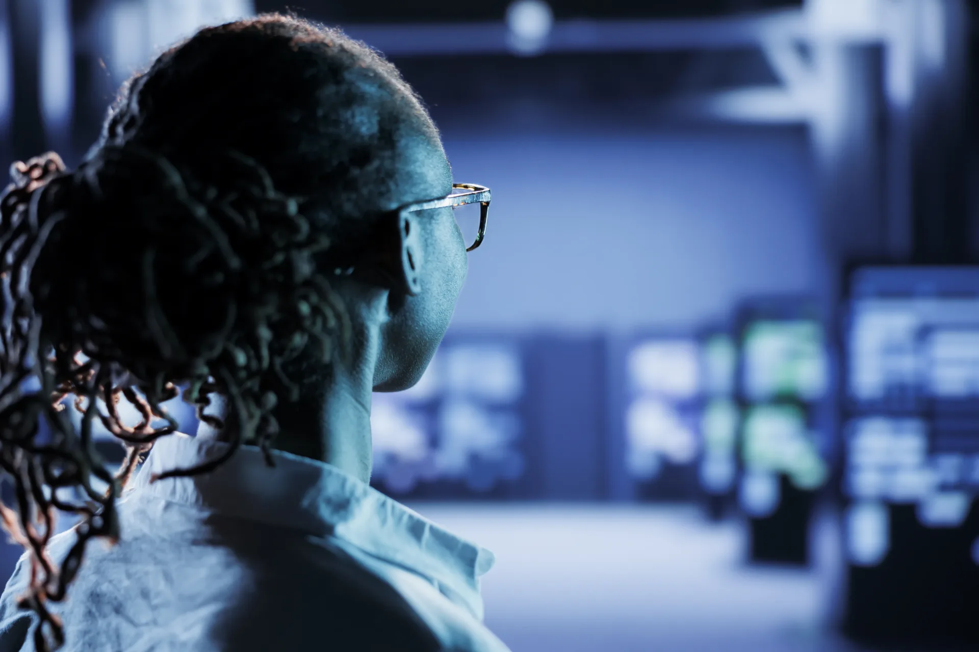 Woman with glasses, seen from behind, looking at data screens in a server room with blue light